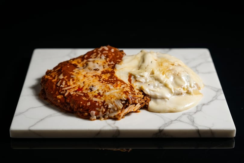 Two enchiladas, one red and one white, on a white marble slab against a dark background.