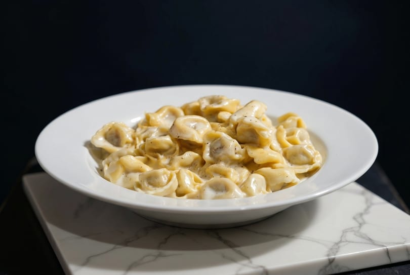 A white bowl filled with creamy tortellini pasta, sprinkled with black pepper, on a marble surface.