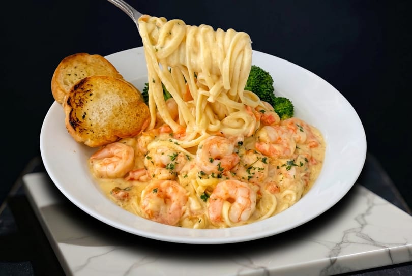 Creamy shrimp linguine with broccoli and garlic bread, lifted by a fork against a dark background.
